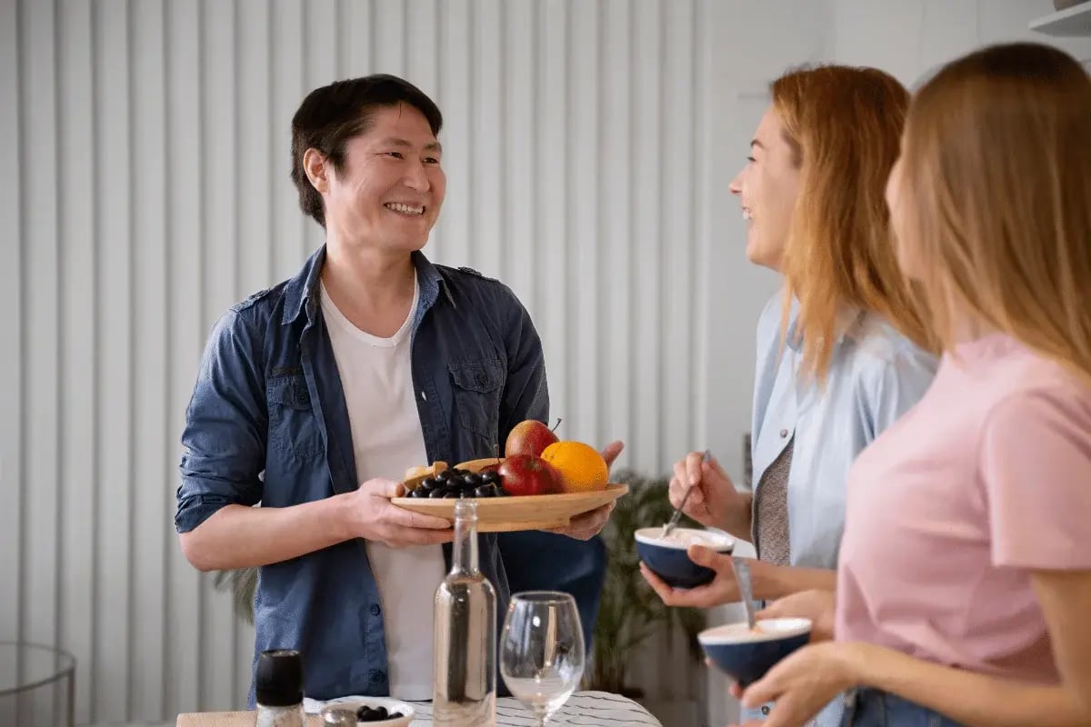 A person holds a wooden platter of fruit, while two others enjoy desserts in a bright, modern kitchen setting.