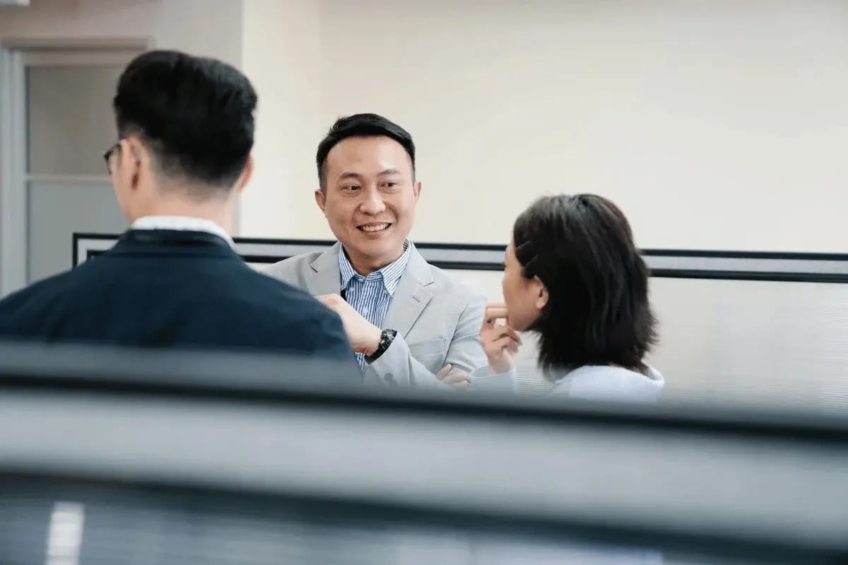 Three professionals having a conversation in an office setting with a focus on a smiling man in a gray suit.