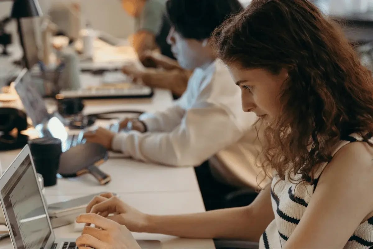 A woman working intently on her laptop at a shared workspace, with a man focused on his computer in the background.