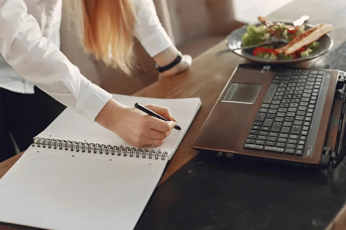 A woman in a white shirt taking notes in a notebook next to a laptop and a bowl of salad.