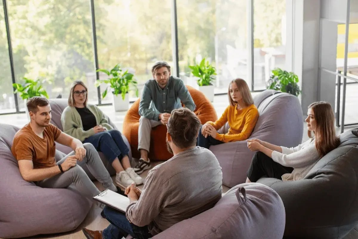 A group of people sit on bean bags in a bright room with large windows and plants, engaged in conversation.