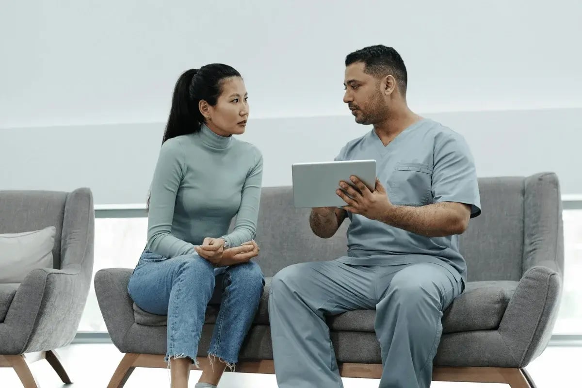A nurse in scrubs shows a digital tablet to a woman in jeans and a sweater. They sit on a couch, engaging in a focused, professional conversation.