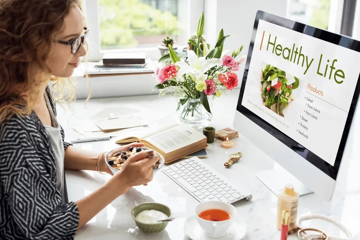 A person enjoys a nutritious meal while working at a desk decorated with flowers, a computer, books, and a cup of tea.