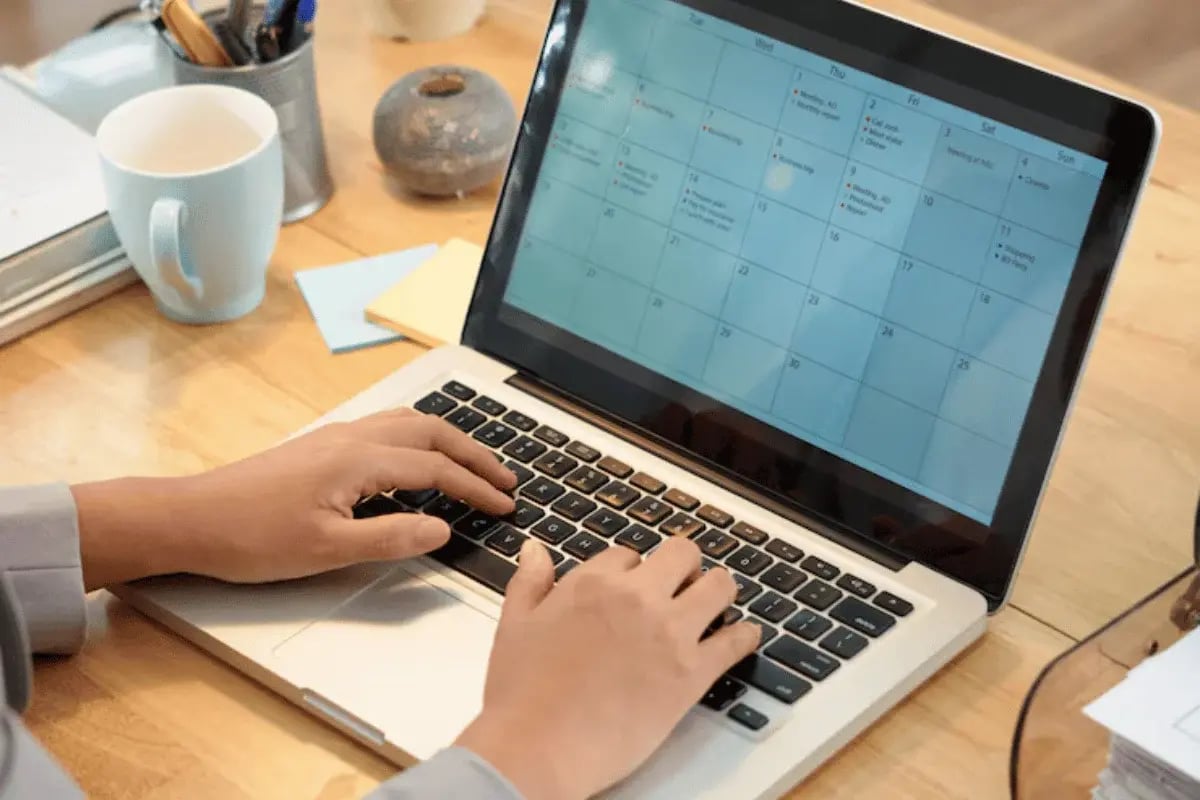 Hands typing on a laptop with a calendar displayed on the screen. The desk is cluttered with a mug, notepad, and stationery, conveying a busy workspace.