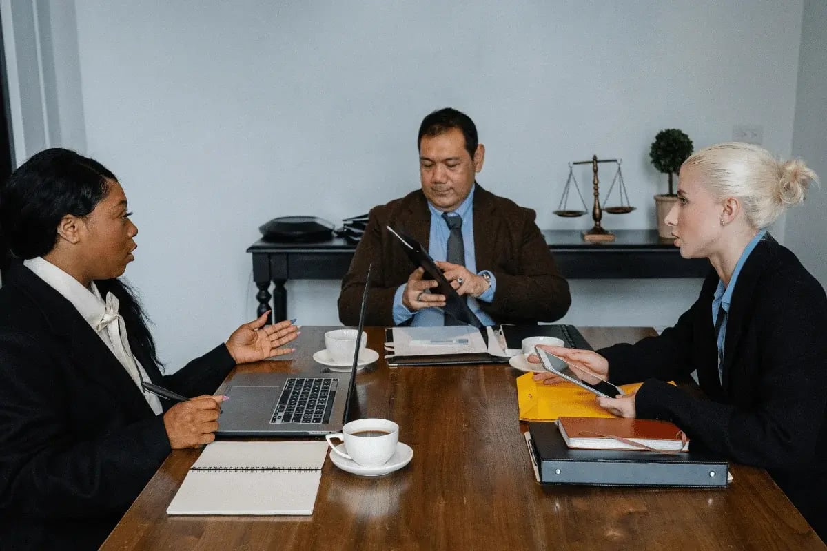 A group of professionals engages in a meeting around a wooden table, with laptops, documents, and coffee cups visible.