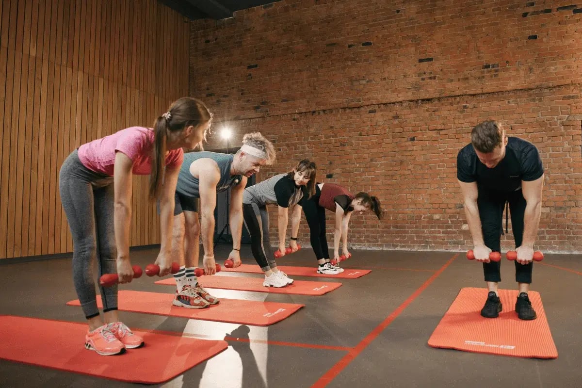 Group fitness class performing dumbbell exercises on red mats in a gym with a brick wall backdrop.