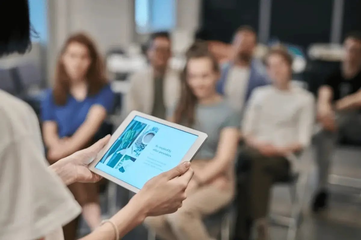 Presenter holding a tablet while giving a presentation to a group of people in a seminar room.