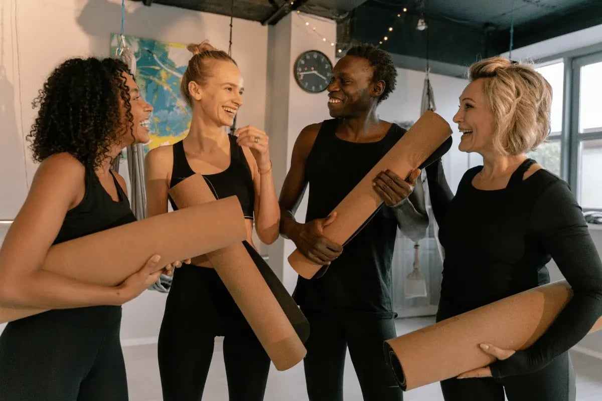 A group of four people smiling and holding yoga mats in a fitness studio.