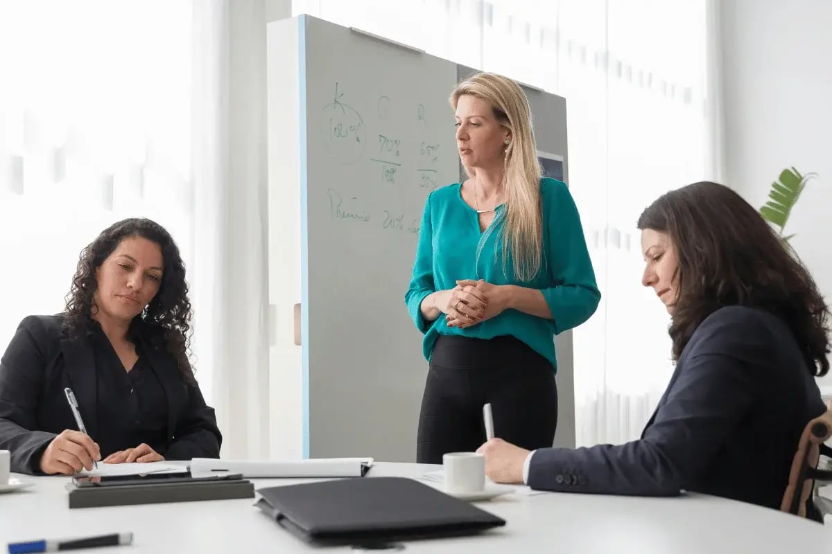 A businesswoman in a teal blouse gestures while presenting ideas to colleagues in a modern office setting.