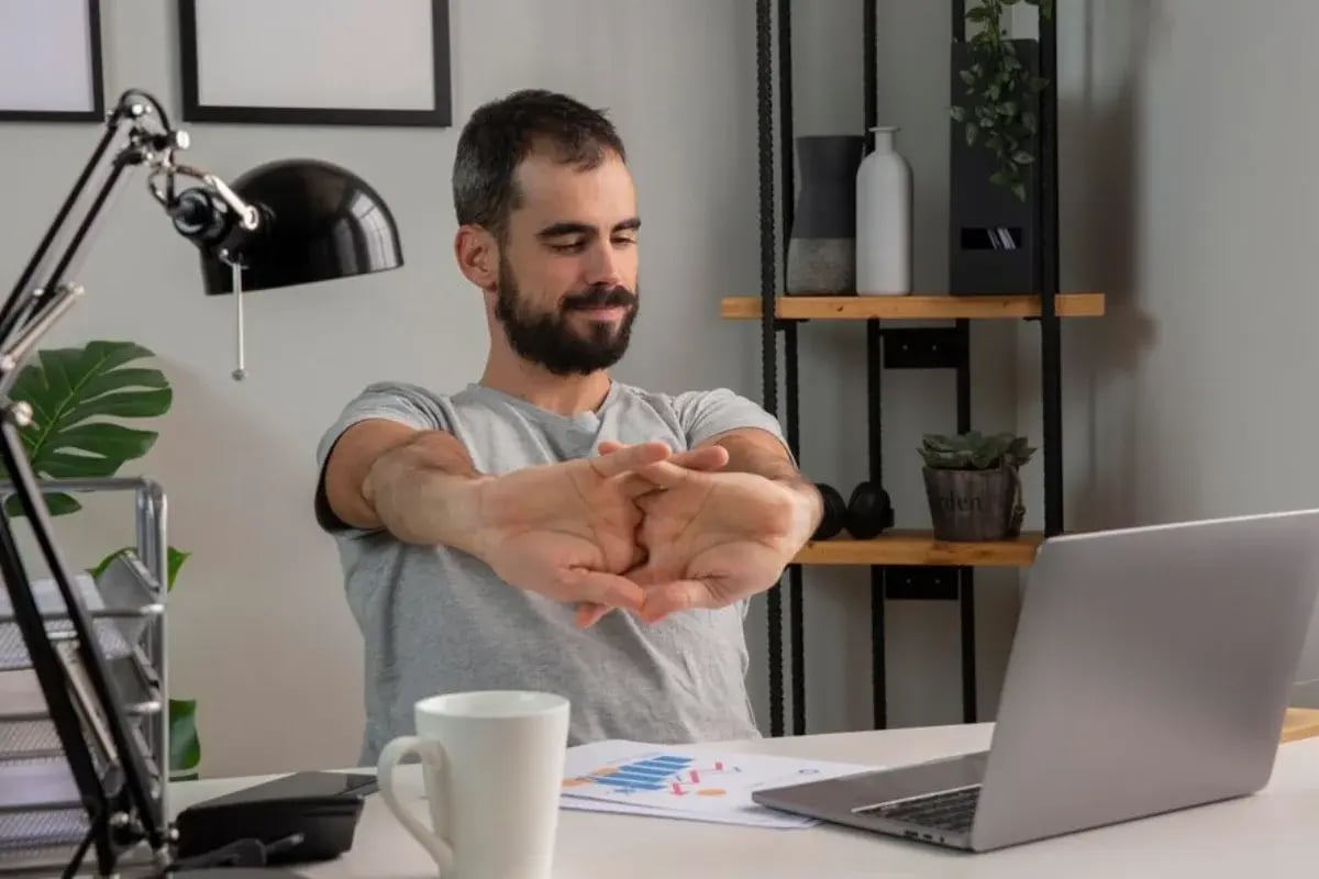 A person stretches their arms while seated at a desk with a laptop, mug, and documents, surrounded by plants and shelves.