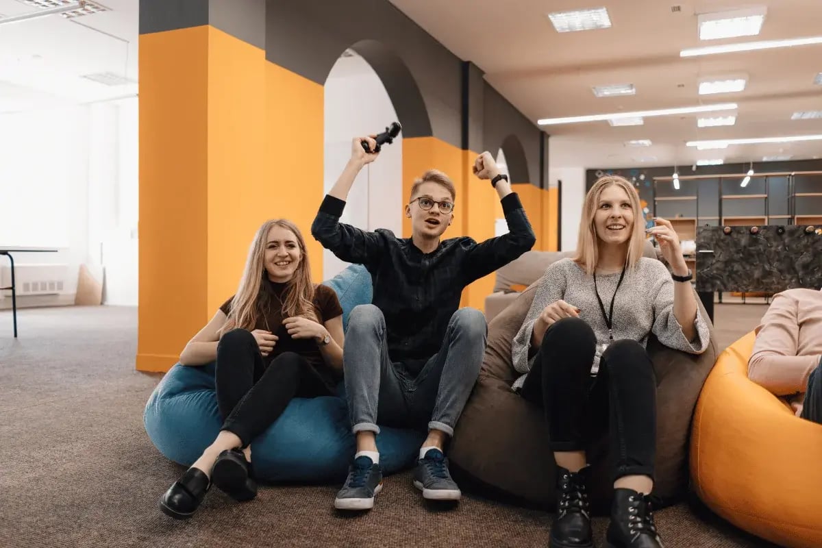 Three people sitting on beanbags, smiling and enjoying a video game session in a bright office space.
