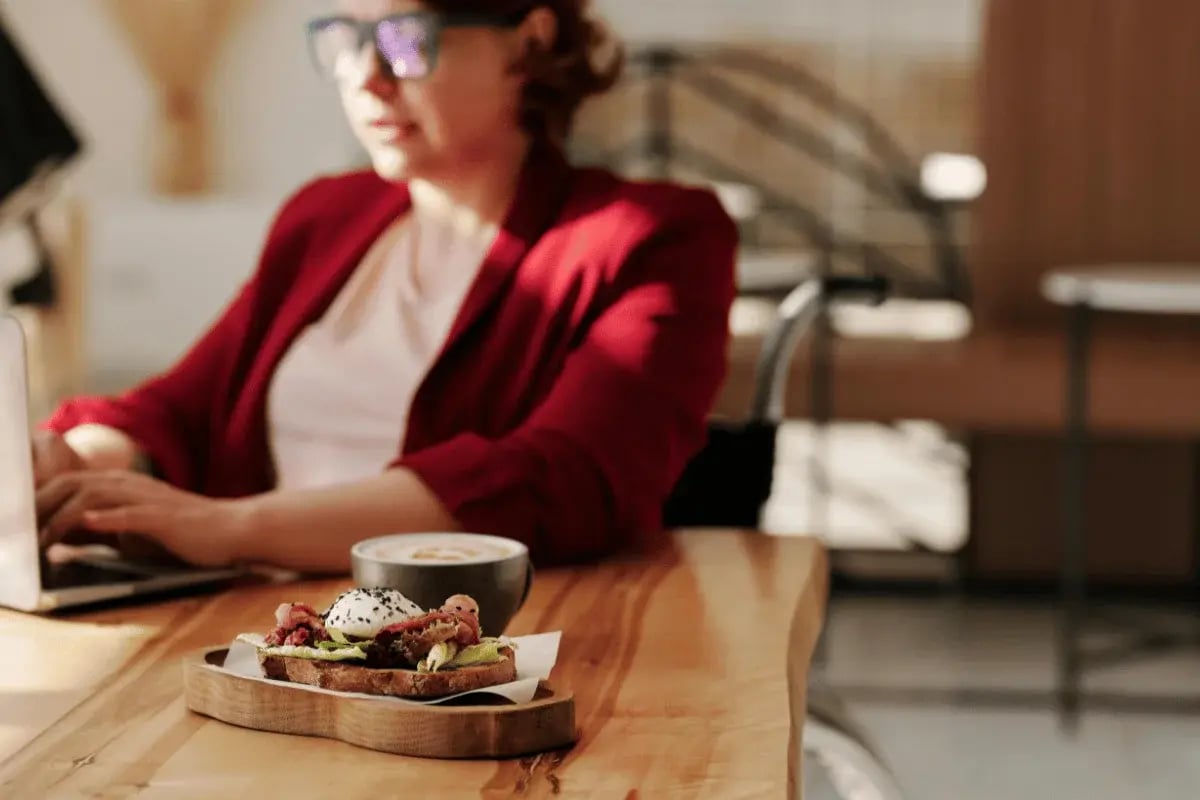 A woman wearing glasses working on a laptop with a delicious breakfast of toast and coffee nearby.
