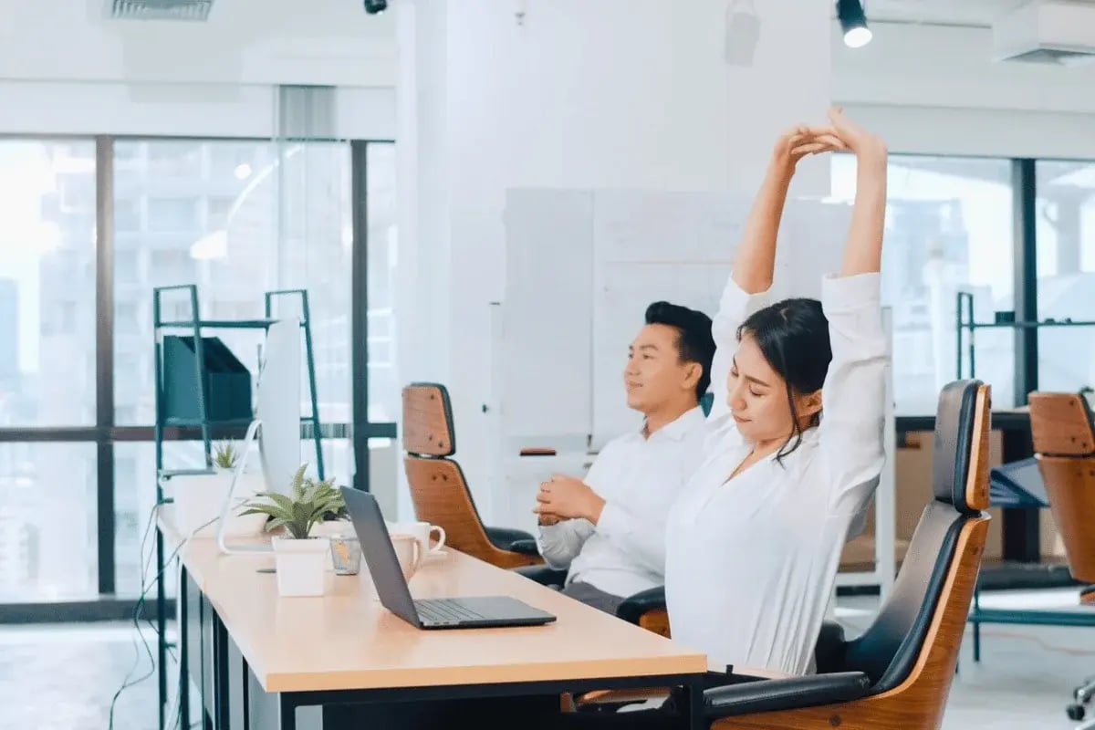 A man and a woman stretching at their desks in an open-plan office with laptops and plants on the desk.