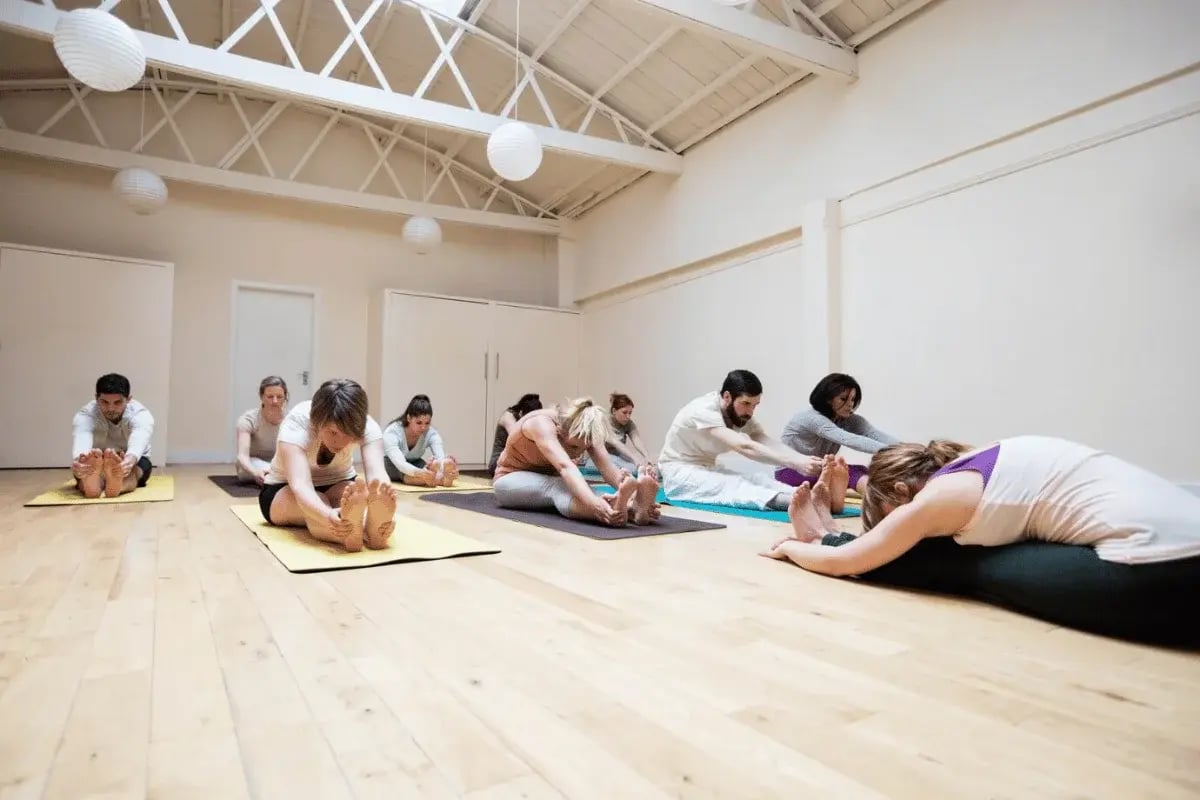 A group of people stretching on yoga mats in a spacious, bright yoga studio with high ceilings.