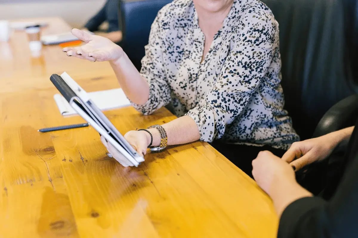 Two people sit at a wooden table, one gesturing while holding a tablet. The scene suggests a business meeting or discussion, conveying collaboration.