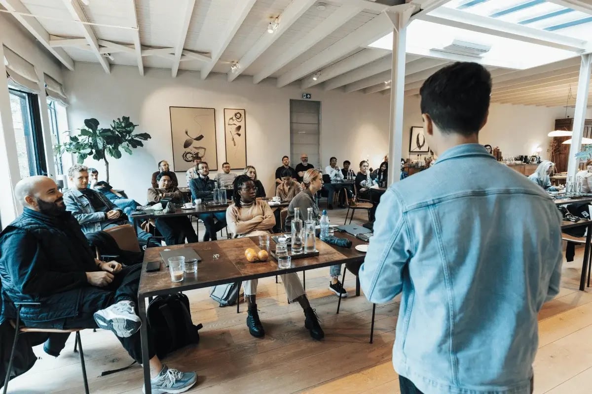 A speaker presenting to a group of people seated at a table in a well-lit office space.