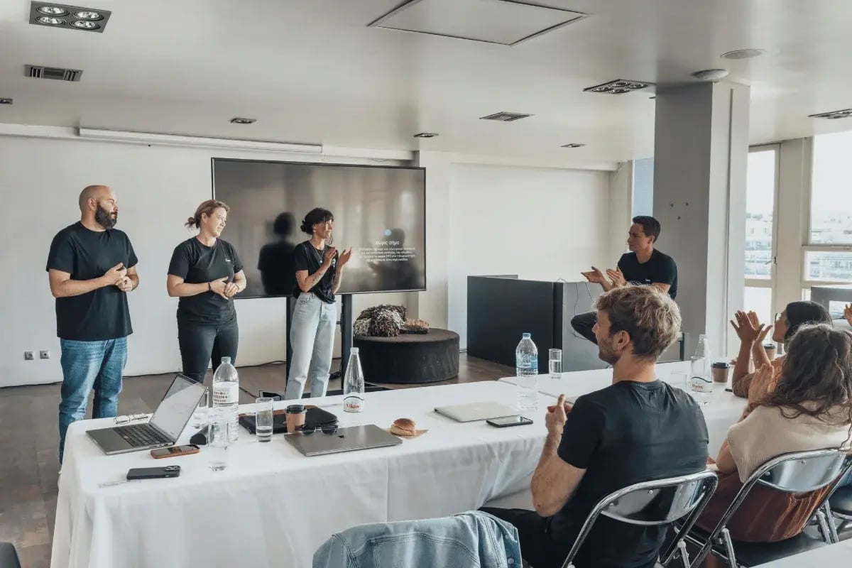 A group of four people stands at the front of a conference room, clapping and smiling, while an audience seated around a table applauds. The atmosphere is collaborative and positive.