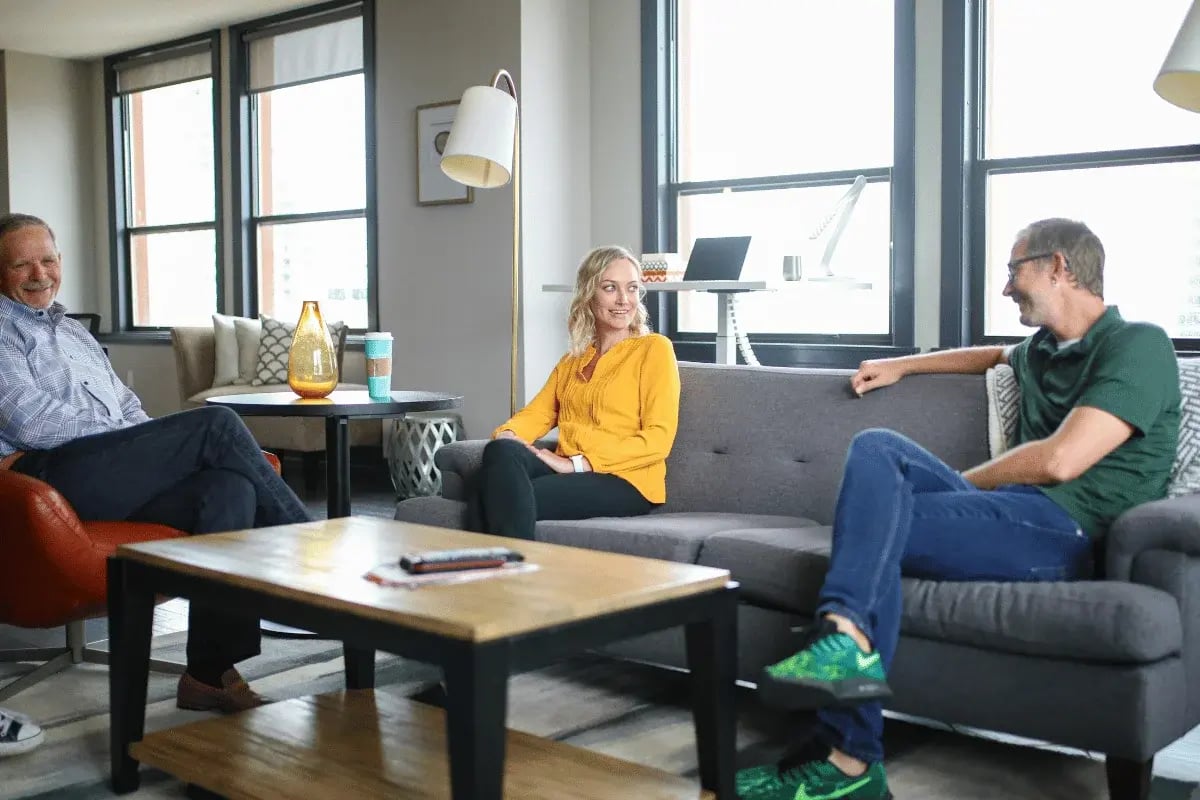 Three people sit in a cozy living room, chatting and smiling warmly. The room features a modern sofa, large windows, and a coffee table creating a relaxed, friendly atmosphere.