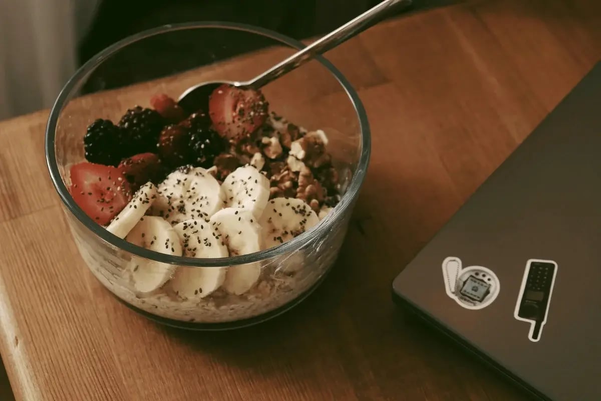 A bowl of granola, banana slices, strawberries, and blackberries beside a laptop on a wooden table.