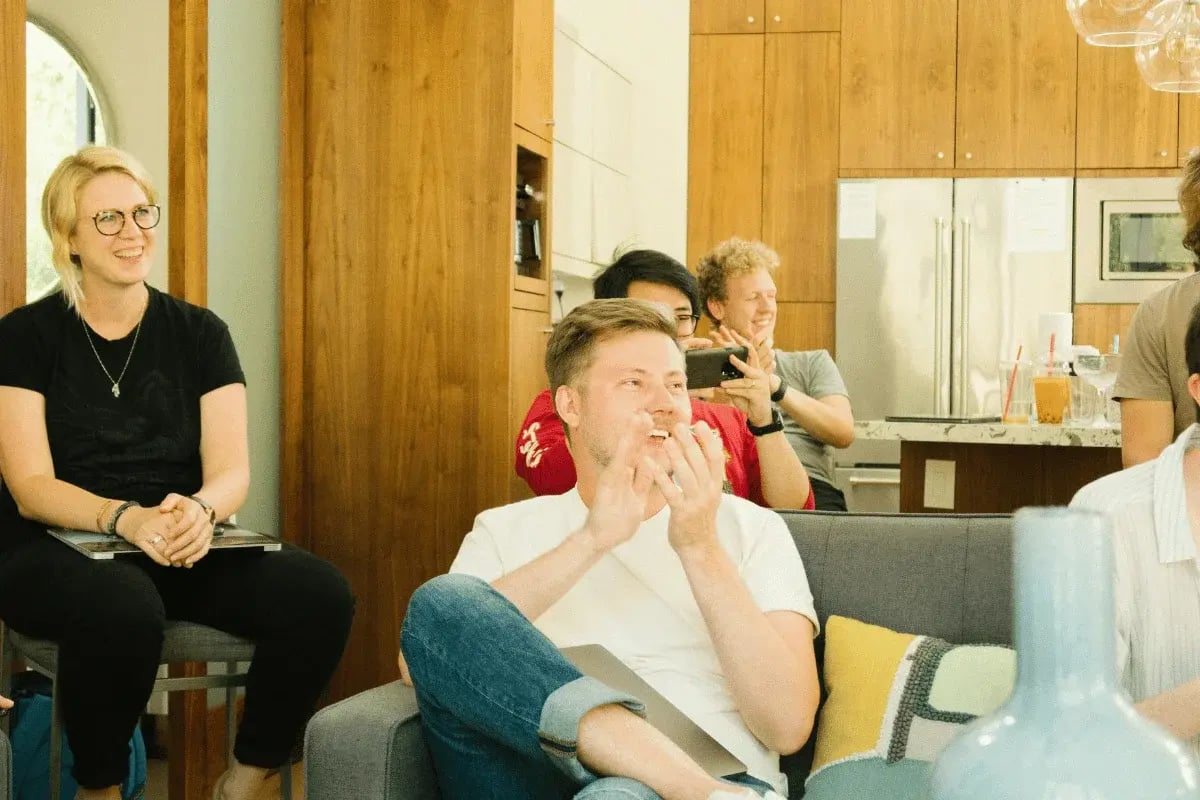 Group of coworkers in a modern kitchen lounge smiling and clapping during a casual office gathering.