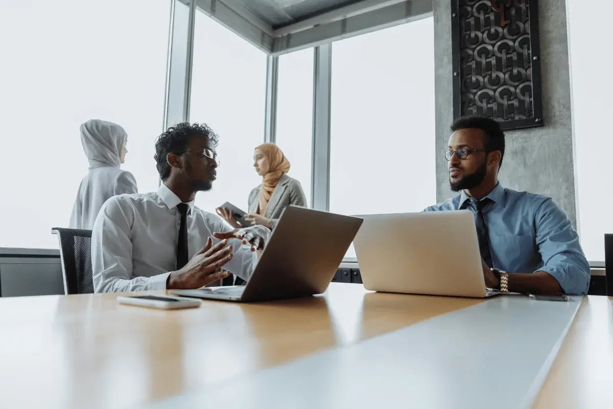 Two men in business attire discuss work at a conference table with laptops. In the background, two women in hijabs engage in conversation by large windows. The scene conveys a professional and collaborative atmosphere.