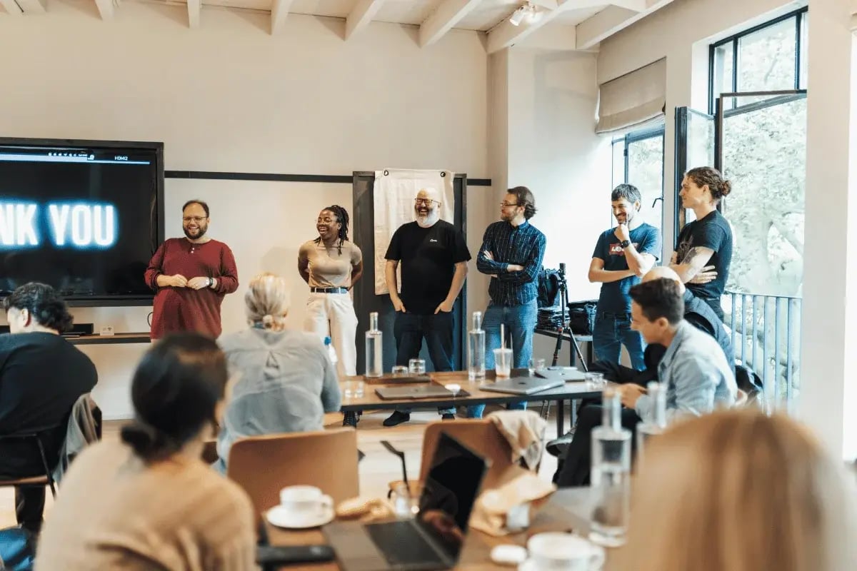 A diverse group of six people stands at the front of a bright, modern room, smiling and engaged. A "Thank You" slide is displayed on a screen beside them. Seated audience members listen attentively, creating an atmosphere of appreciation and collaboration.