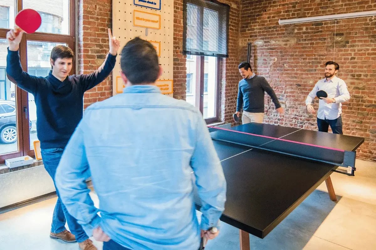 A group of men playing ping pong in an office, one celebrating a point while others prepare to return the ball.