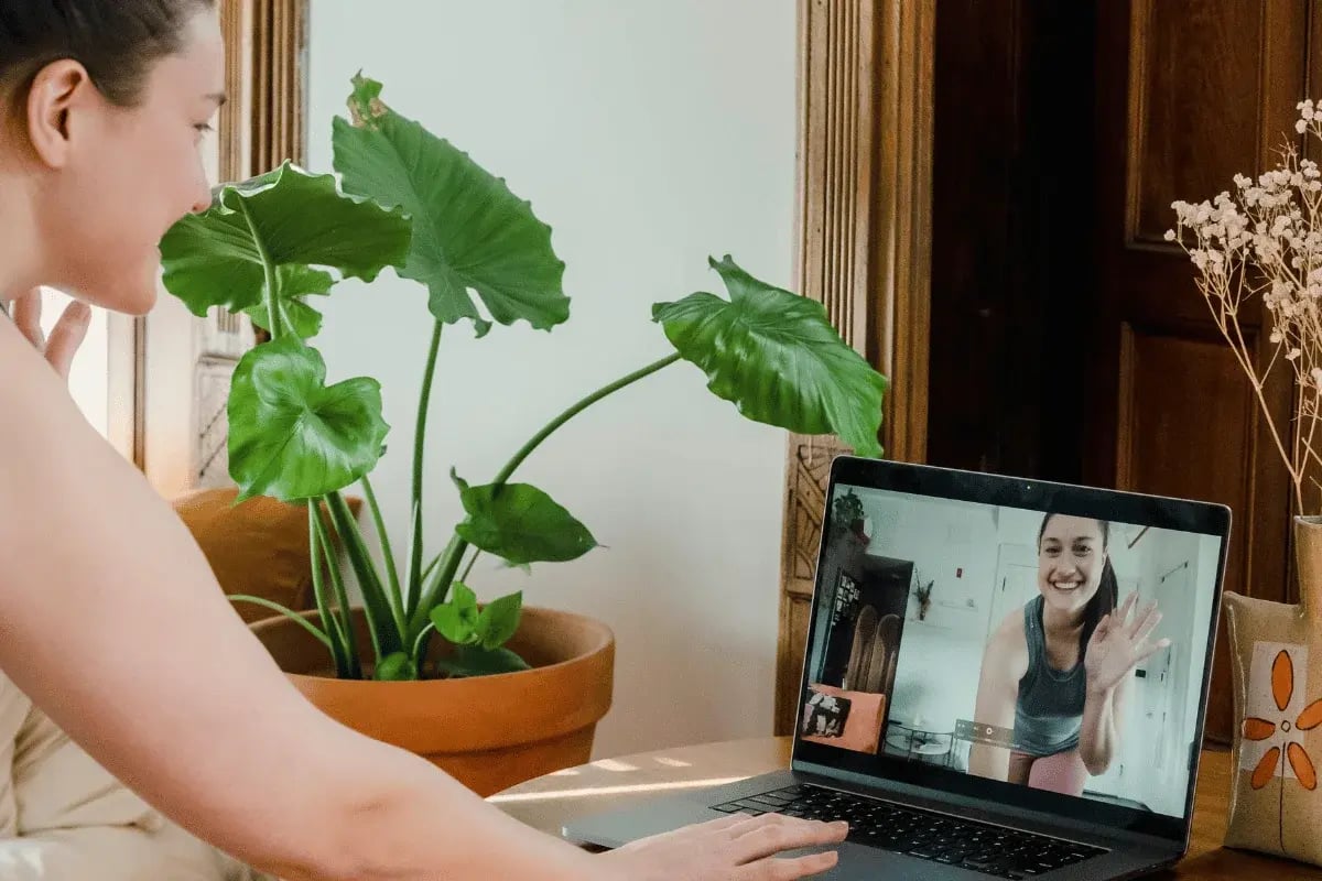 A person interacting via video call on a laptop, with a large green plant in the background and a cozy home setting.