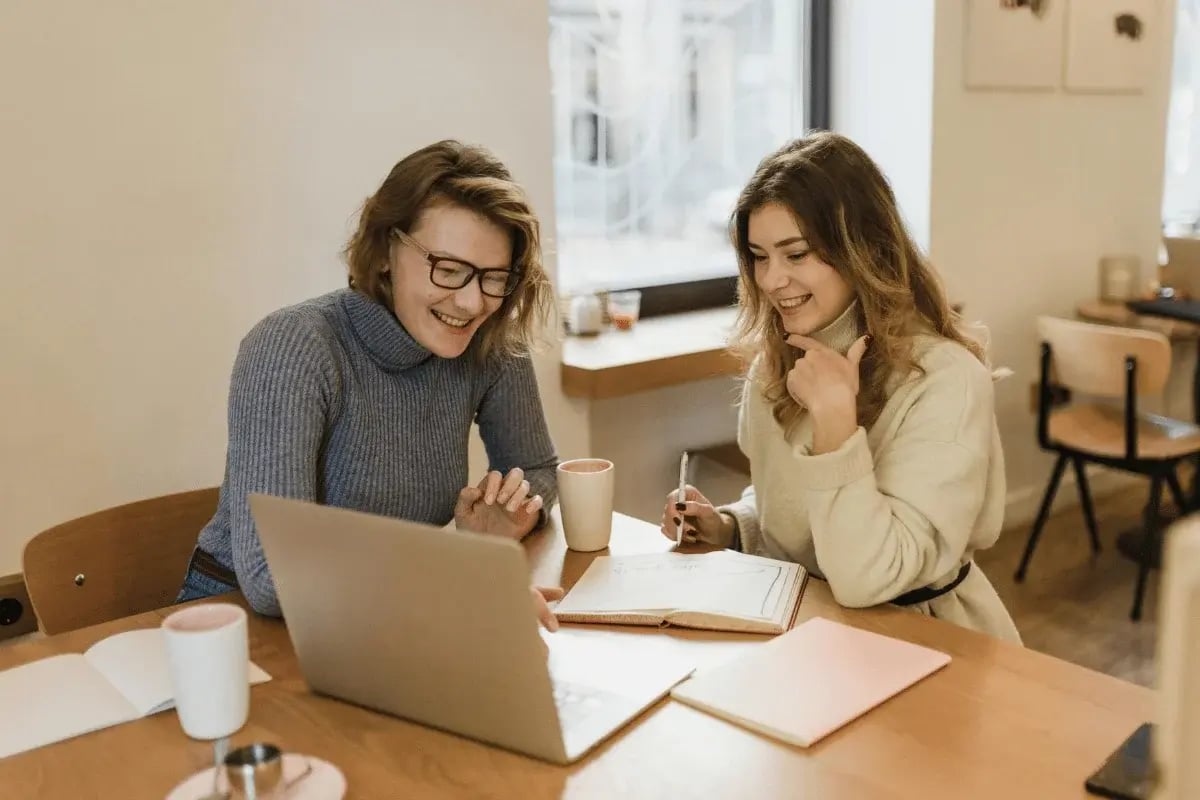 Two women smiling while working on a laptop in a cozy café.