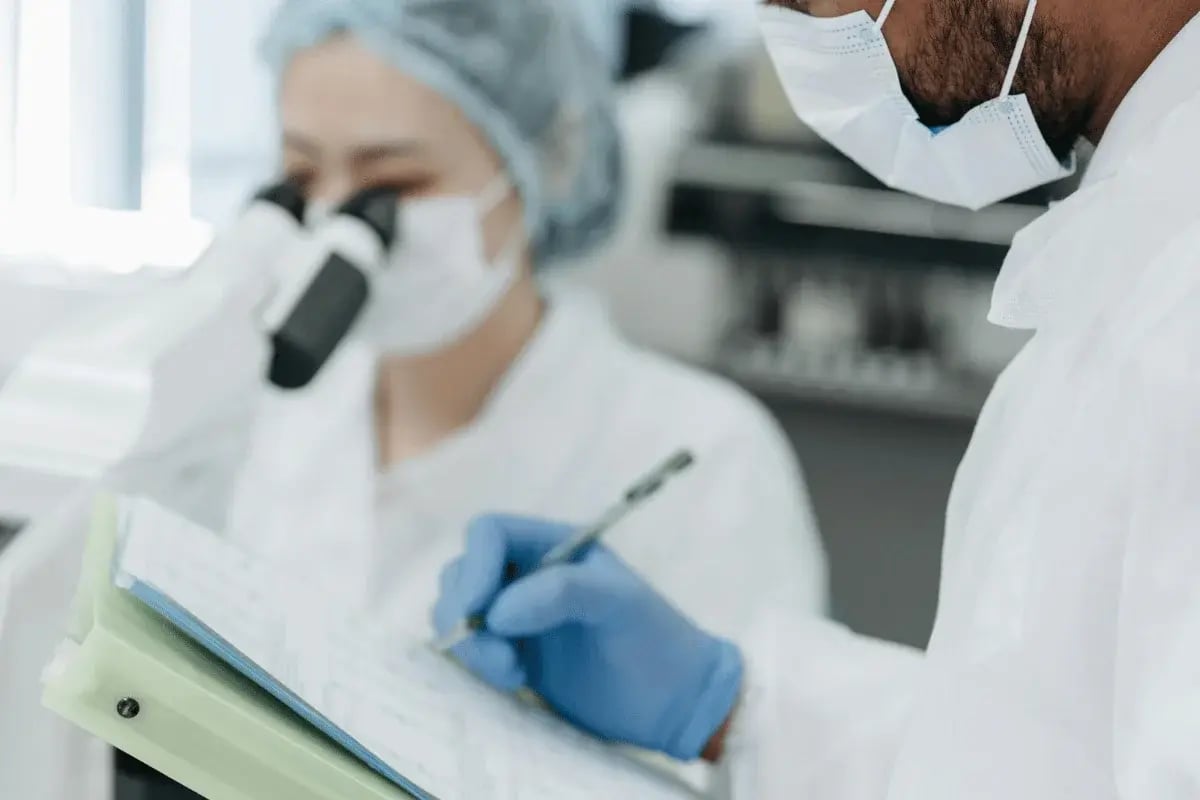 Two scientists in lab coats and masks work in a lab. One focuses on a microscope, while the other writes notes on a clipboard, wearing blue gloves.