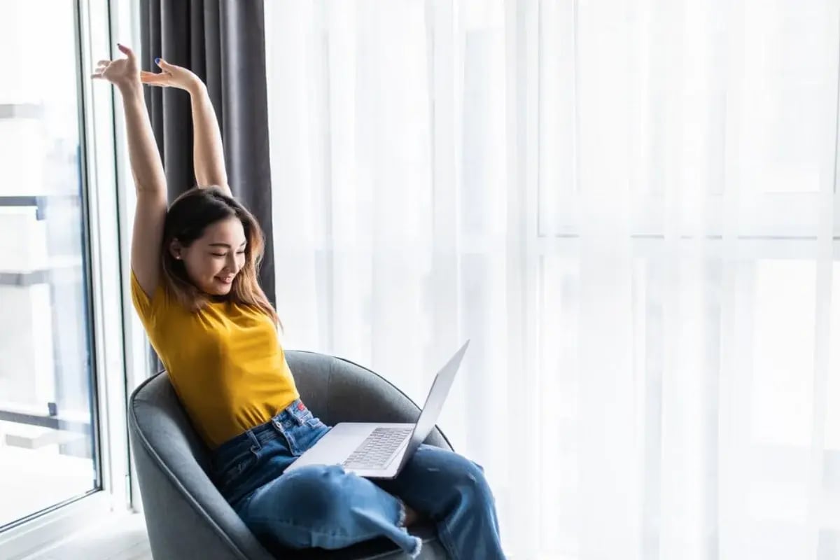 A person in a yellow shirt stretches while sitting in a gray chair, using a laptop near a large window with sheer curtains.
