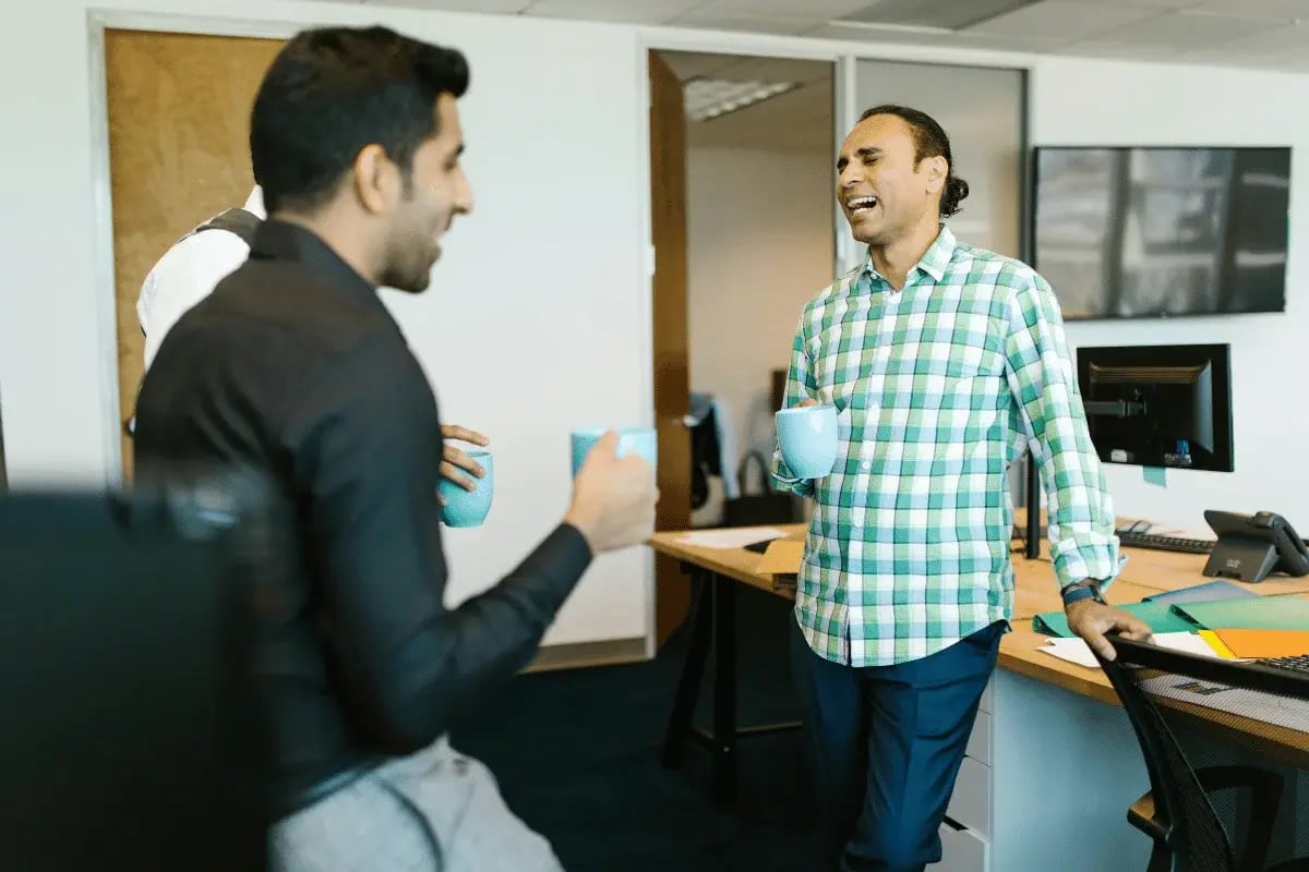 Office coworkers sharing a laugh and coffee during a break in a modern open-plan workspace.