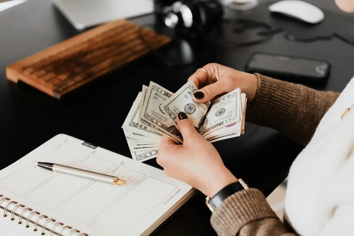 Woman counting a stack of US dollar bills next to a planner and pen on a black office desk