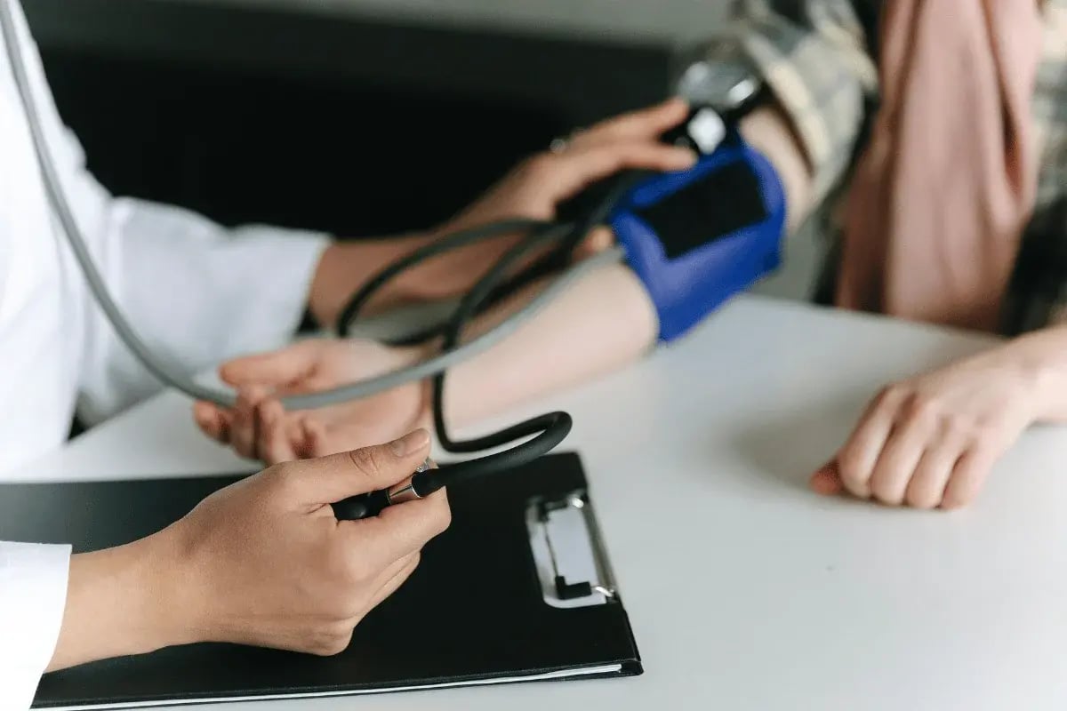 A person in a white coat uses a stethoscope and blood pressure cuff on another person's arm. A clipboard rests on the table. The scene is calm and professional.