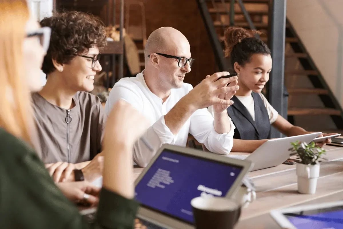 A diverse group of professionals engaged in a collaborative meeting, discussing ideas with laptops and a notebook on a wooden table.