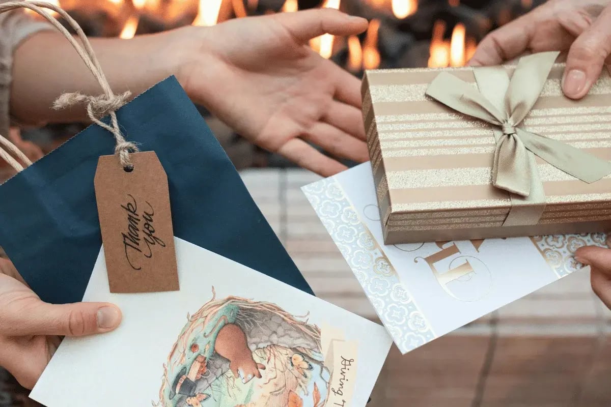 Two hands exchange a decorated gift box and colorful cards, including one with a "Thank You" tag, against a warm fireplace backdrop.