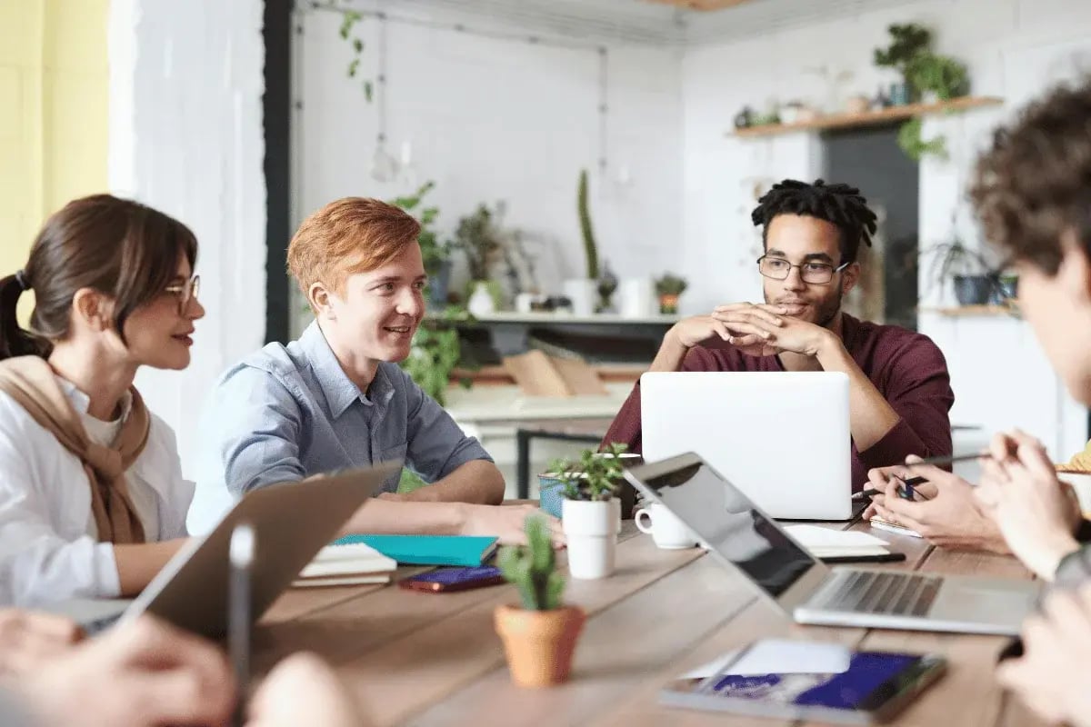 A diverse group of young professionals engage in a meeting around a wooden table, with laptops and notebooks.