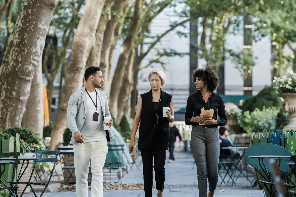 Three professionals walking through a tree-lined outdoor plaza with takeaway coffee and snacks in hand.
