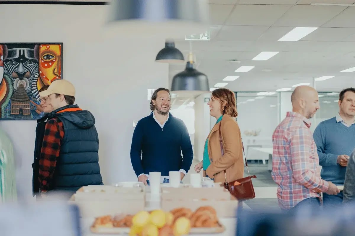 A group of people casually chat in a bright office setting. A colorful abstract painting decorates the wall. The atmosphere is relaxed and social.