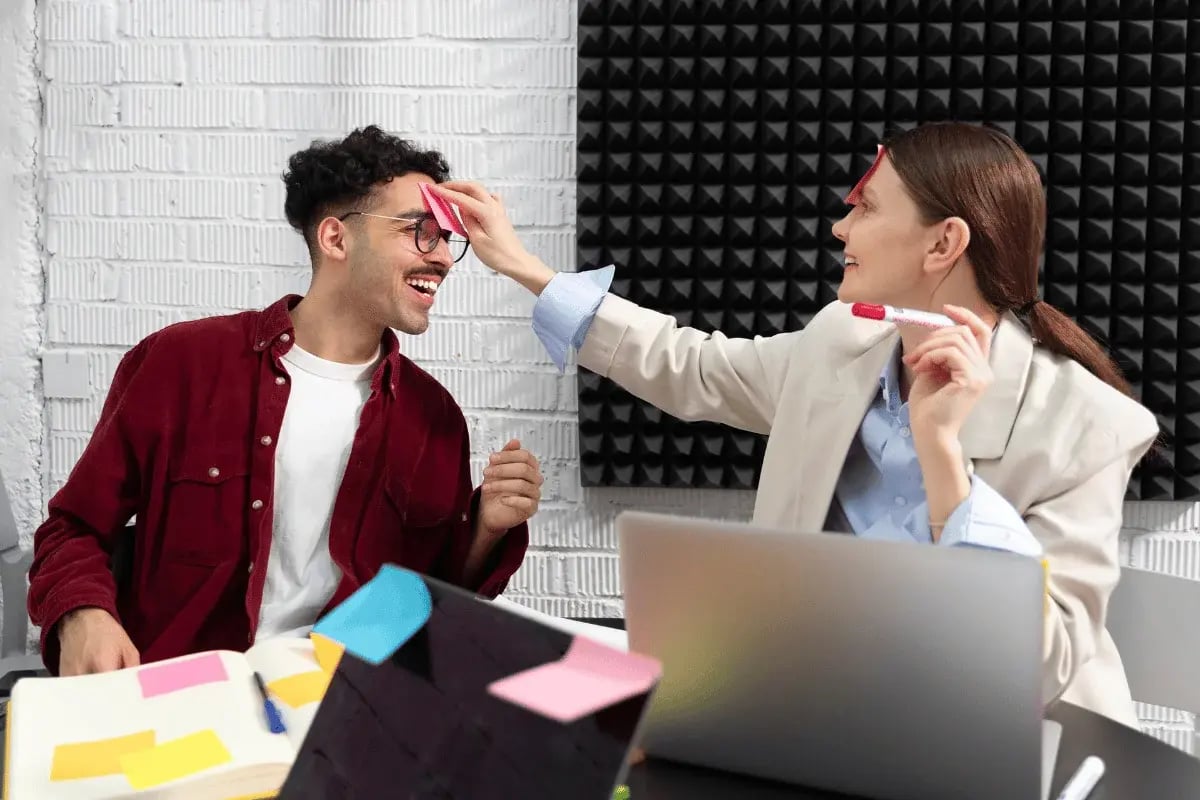 Two coworkers laughing and placing sticky notes on each other’s foreheads.