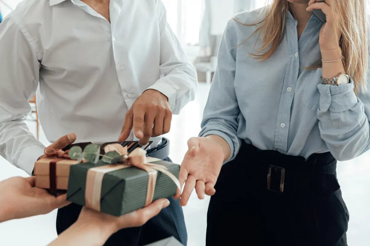 A man hands a gift wrapped in green and brown paper to a woman, while another woman looks on, holding a phone to her ear.