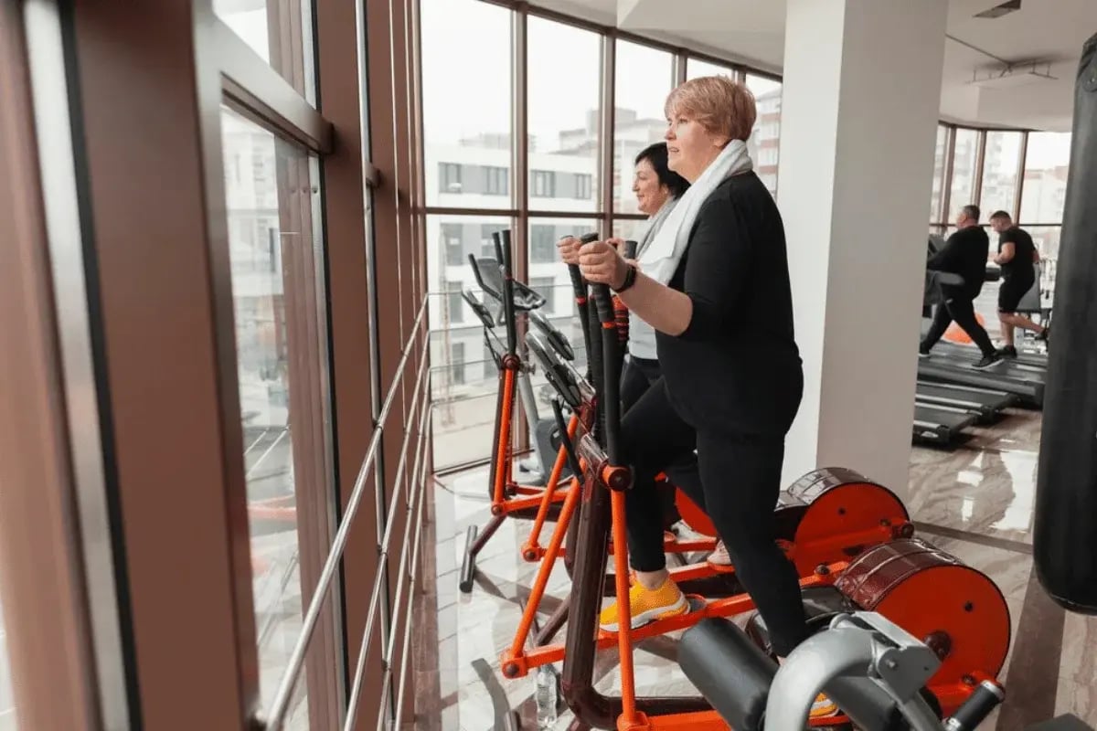 Two women using ellipticals in a gym while looking out the large windows.