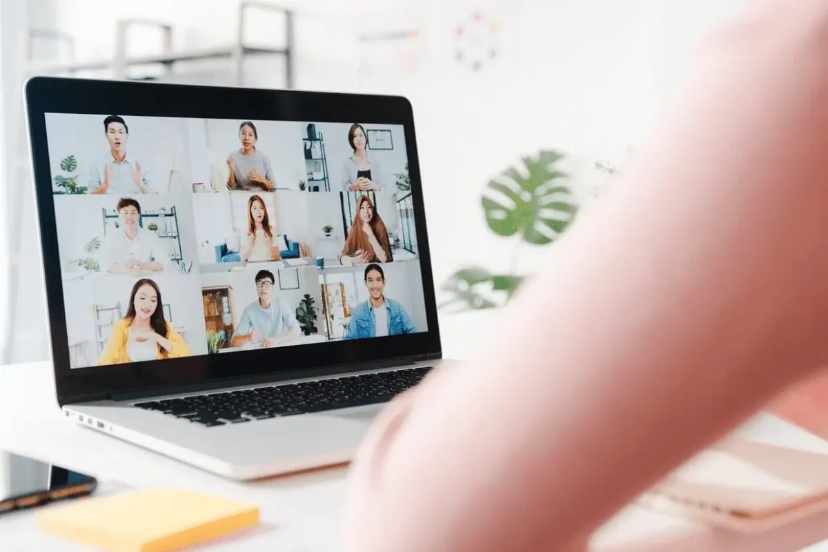 A laptop screen displays a video call with multiple participants, set in a bright, modern workspace with greenery in the background.