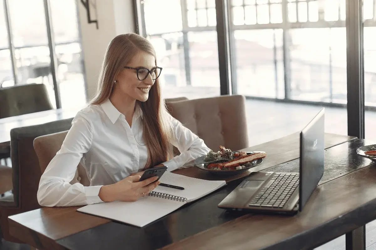 A woman with long hair and glasses smiles while holding a phone, facing a laptop in a bright cafe. Open notebook and plate of food on the wooden table.