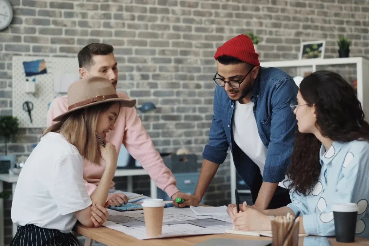 A diverse group of four casually dressed individuals collaborates around a table with blueprints in an office with a brick wall. They appear focused and engaged.