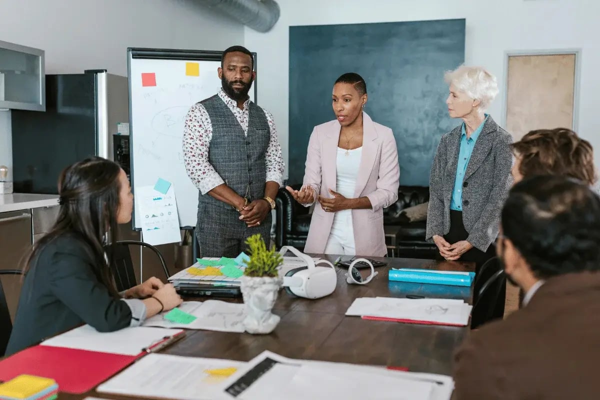 A diverse group of professionals discusses around a table in an office. A whiteboard and virtual reality headset are visible, suggesting a creative meeting.