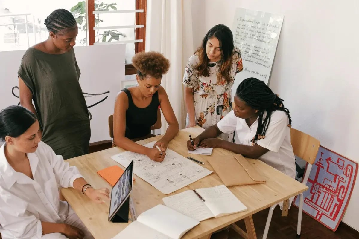 Five women collaborate around a table with papers and a laptop, conveying teamwork and focus in a bright room with notes on a board.