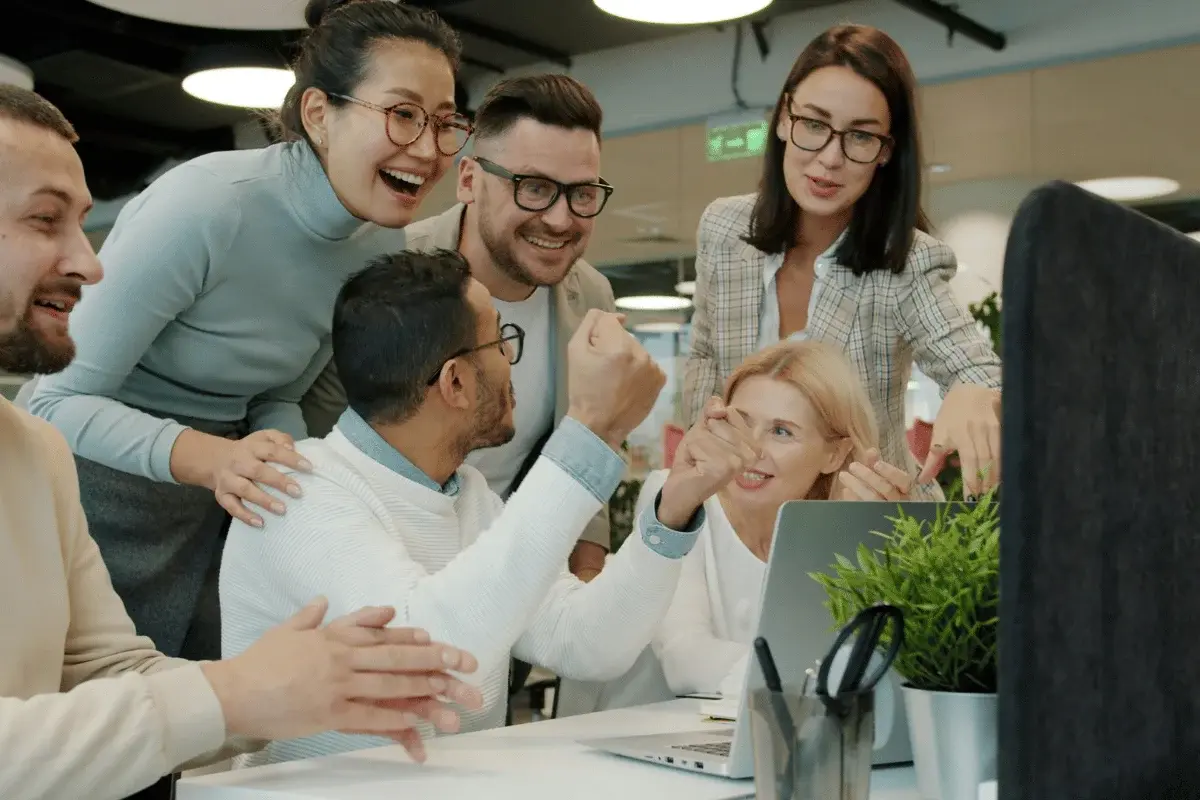 A group of people in a modern office setting, engaged in an animated discussion around a laptop, displaying excitement and collaboration.