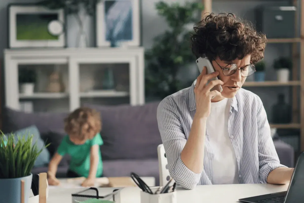 A person is on the phone at a desk while a child in a green shirt draws nearby. The setting is a cozy, modern living room.