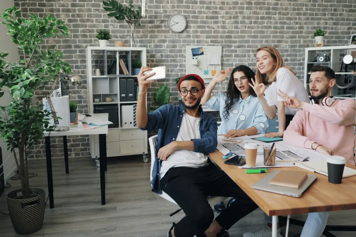 A diverse group of four smiling people take a selfie in a modern office with greenery and bookshelves, conveying a casual and creative atmosphere.